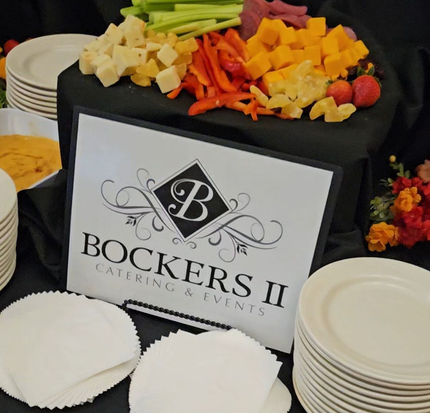 Catering table with cheese, veggies, and a sign for Bockers II Catering & Events, with stacks of white plates.