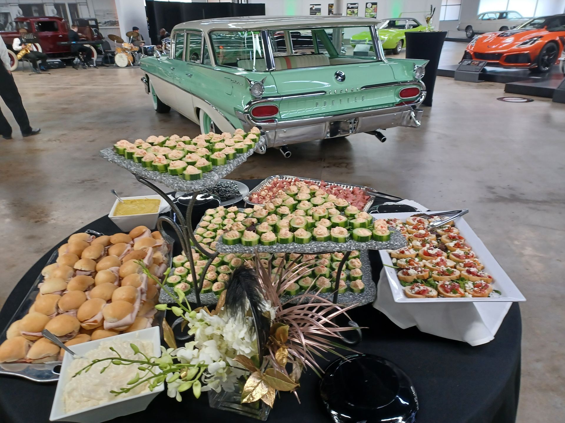 A buffet table with appetizers and sandwiches in a spacious hall with several vintage and modern cars in the background.