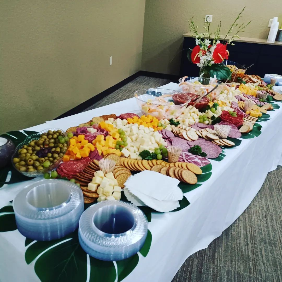 A long buffet table covered with a white cloth, filled with platters of cheese, meats, crackers, olives, and flowers.
