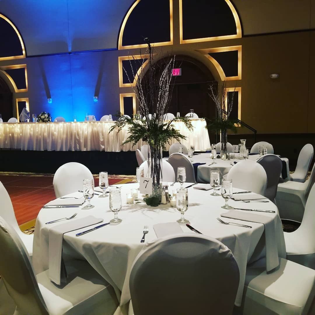 A round wedding reception table with white linens, centerpieces, and chairs in a banquet hall with blue accent lighting.