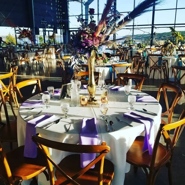 A decorated round wedding table with lavender napkins and a tall floral centerpiece in a bright, modern event venue.