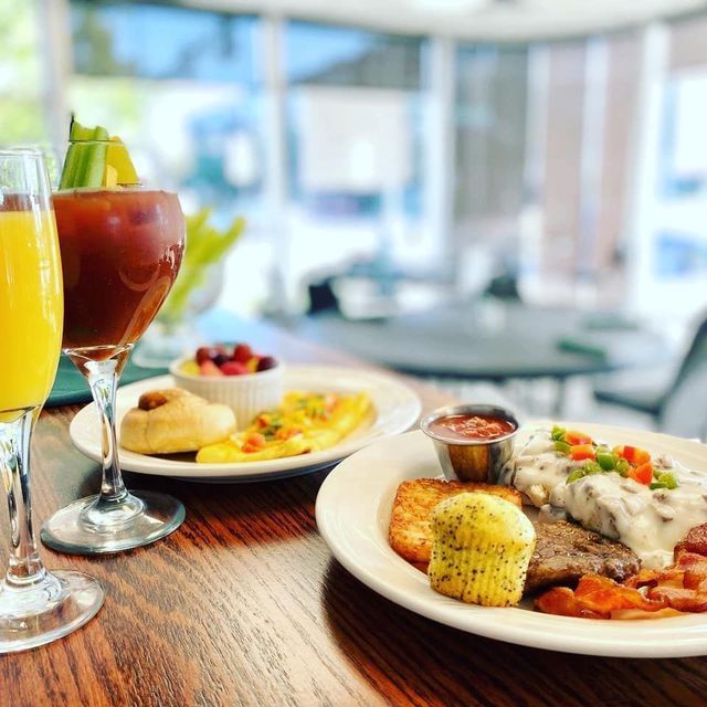 A breakfast spread with two plates of food, a mimosa, and a Bloody Mary on a wooden table in a bright restaurant.