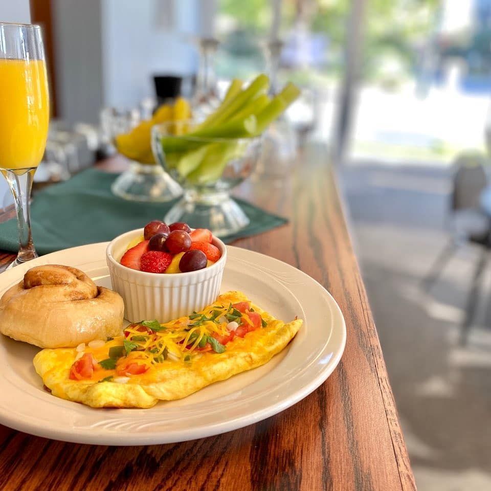 A breakfast plate featuring an omelet, fruit cup, and a pastry, with a mimosa and celery sticks nearby on a wooden table.