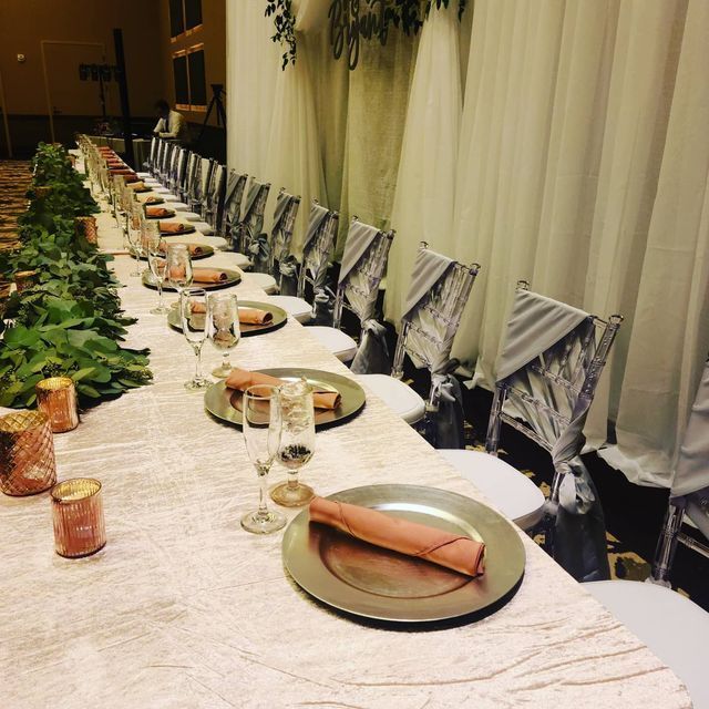 A long banquet table set with metallic chargers, pink napkins, and greenery against draped curtains and decorated chairs.