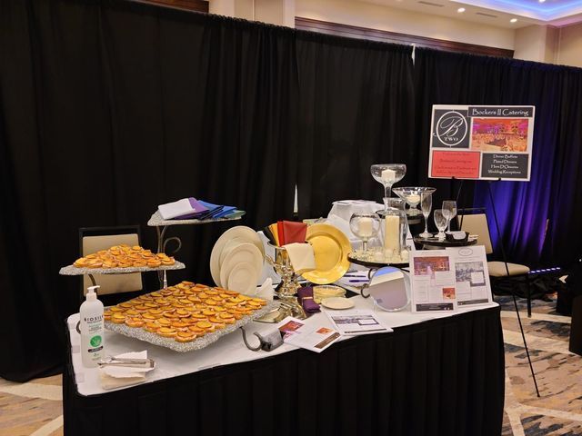 A catering display with appetizers, stacks of white and gold plates, and informational signs on a black-draped table.
