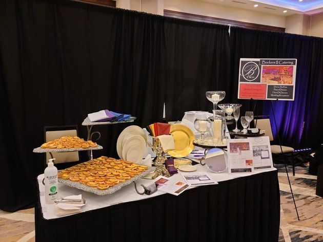 Catering display table with trays of appetizers, stacked plates, glassware, and business signs against a black backdrop.