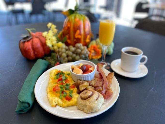 A breakfast plate with eggs, bacon, fruit, and a cinnamon roll, alongside coffee, orange juice, and fall decorations.