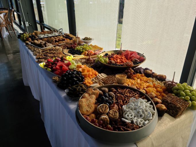 A long buffet table displays several bowls and arrangements of fruits, crackers, cheeses, and snacks in a bright room.