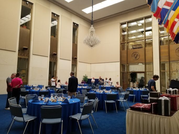 People gather in a high-ceilinged hall for an event, with round blue-covered tables and a draped table of gifts.