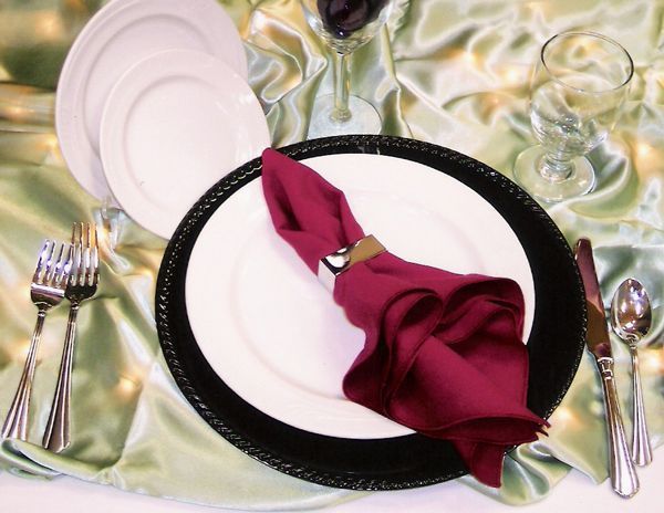 A formal place setting on a sage green tablecloth, featuring a black charger, white plates, a magenta napkin, and silverware.