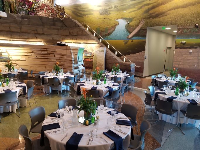 A banquet room with round tables, white tablecloths, dark napkins, and floral centerpieces under a large landscape mural.