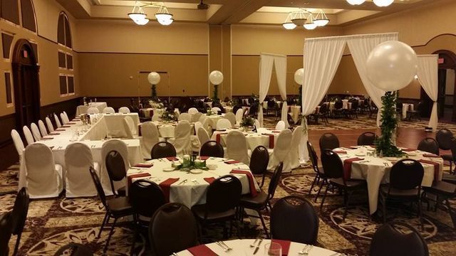 A banquet hall set for a wedding reception with round tables, white tablecloths, maroon runners, and large white balloons.