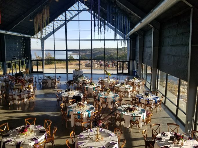 A high-angle view of a bright event hall with round tables, chairs, and large glass walls overlooking a lake.