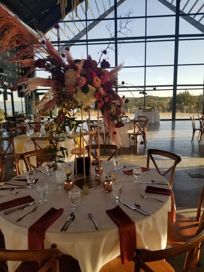 A formal round dining table set for an event inside a large glass-walled venue with a tall, vibrant floral centerpiece.