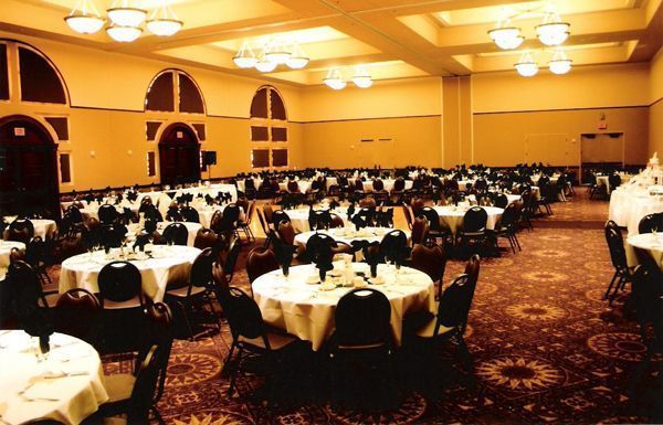 A large, dimly lit banquet hall filled with round tables covered in white tablecloths and dark chairs under ceiling lights.