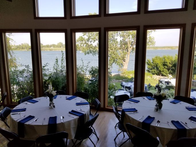 Two round dining tables with white tablecloths and navy napkins, set before large windows overlooking a lake.