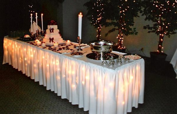 A long table set for a wedding, decorated with a white skirt, string lights, a tiered cake, and silver serving dishes.