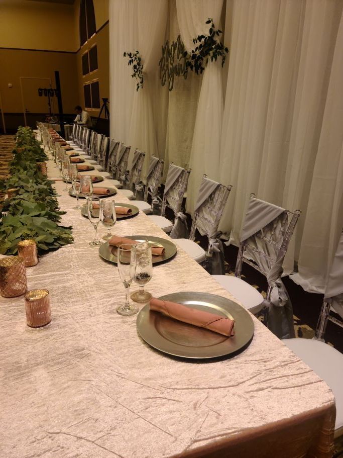 A long banquet table set for a formal event with pink linens, metallic chargers, champagne flutes, and floral greenery.