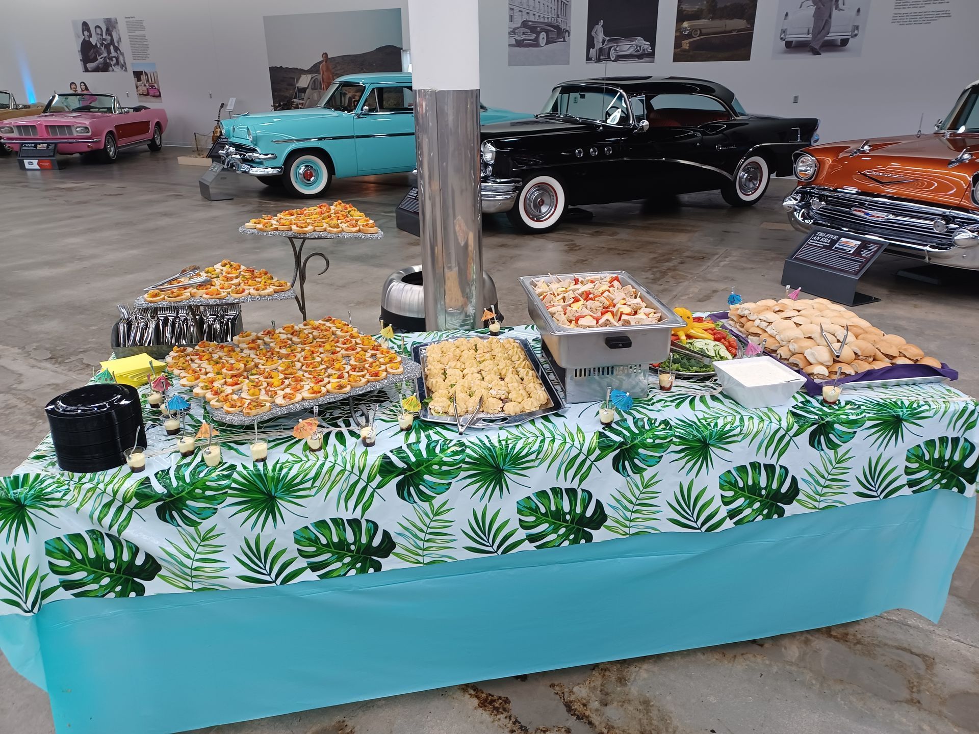 A food buffet table with snacks and pastries, draped in a tropical print tablecloth, set inside a classic car museum.
