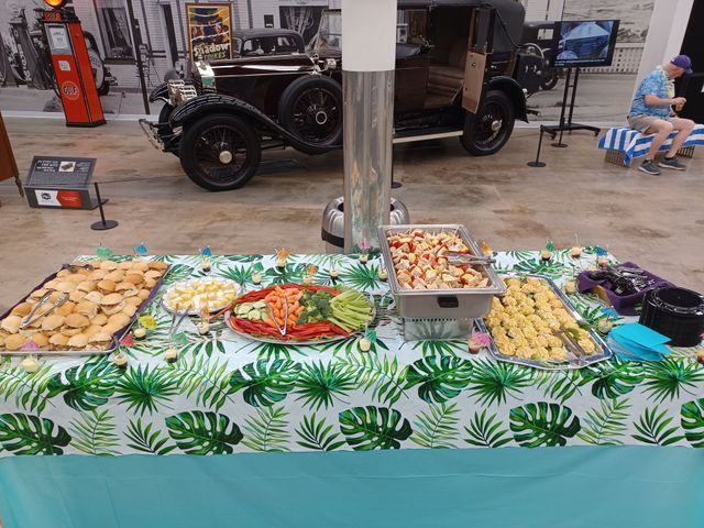 A food buffet table with snacks and fruit at a museum, positioned in front of a vintage car display.