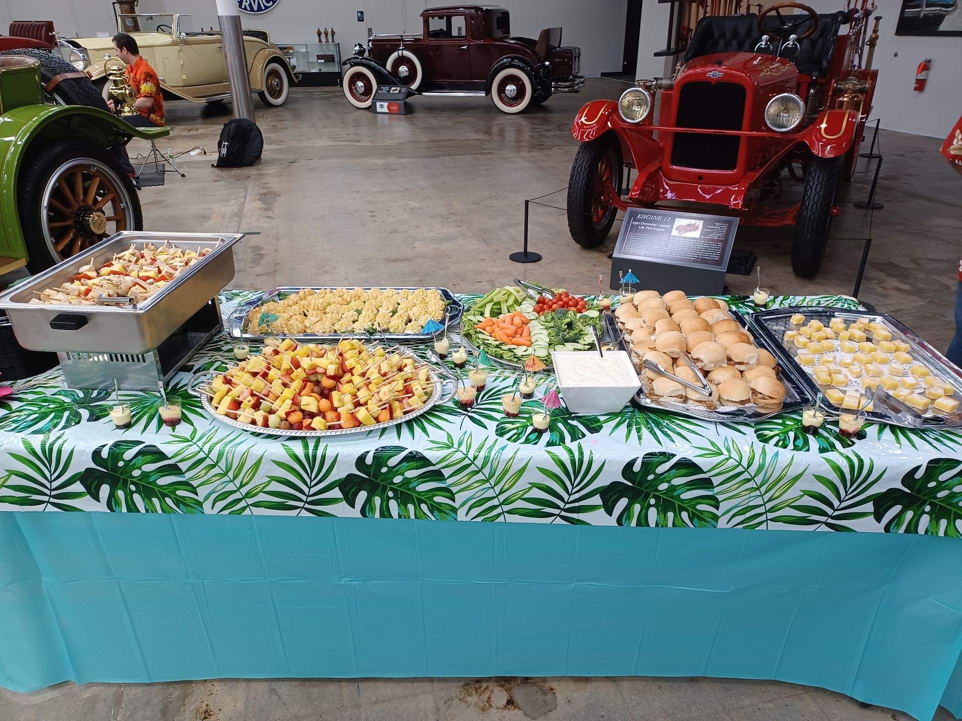 Buffet table with assorted appetizers and rolls, arranged on a palm leaf patterned cloth inside a vintage car museum.