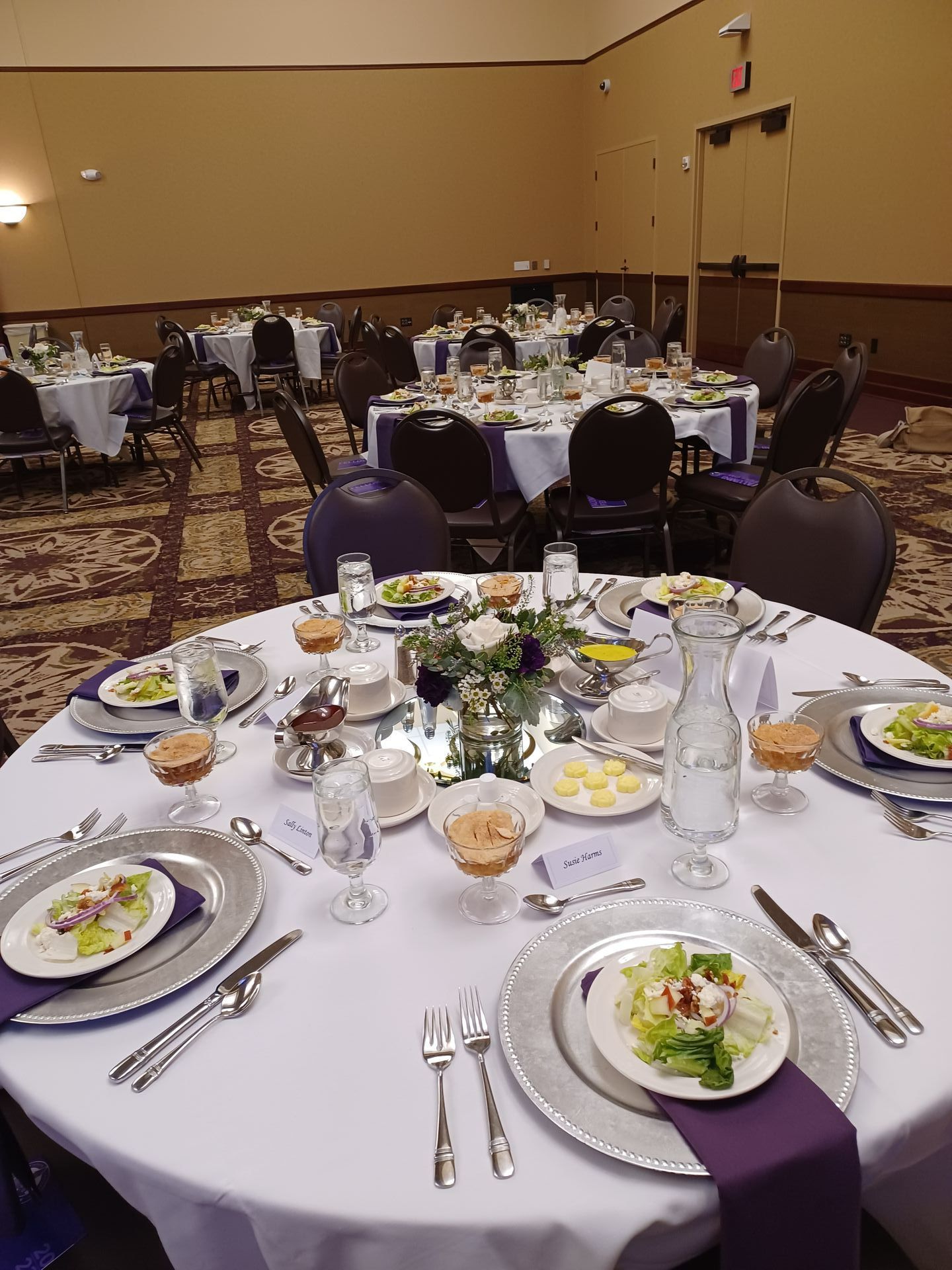 A banquet hall with many round tables set for a dinner, featuring white tablecloths, purple napkins, and salad plates.