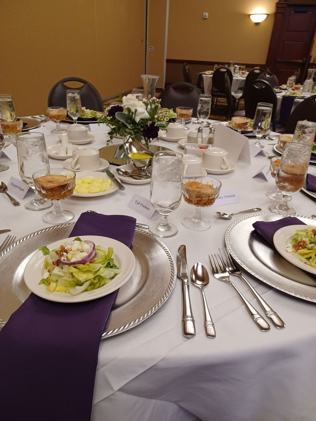 A formal dining table set for an event with purple napkins, silver charger plates, glasses, and salad plates.