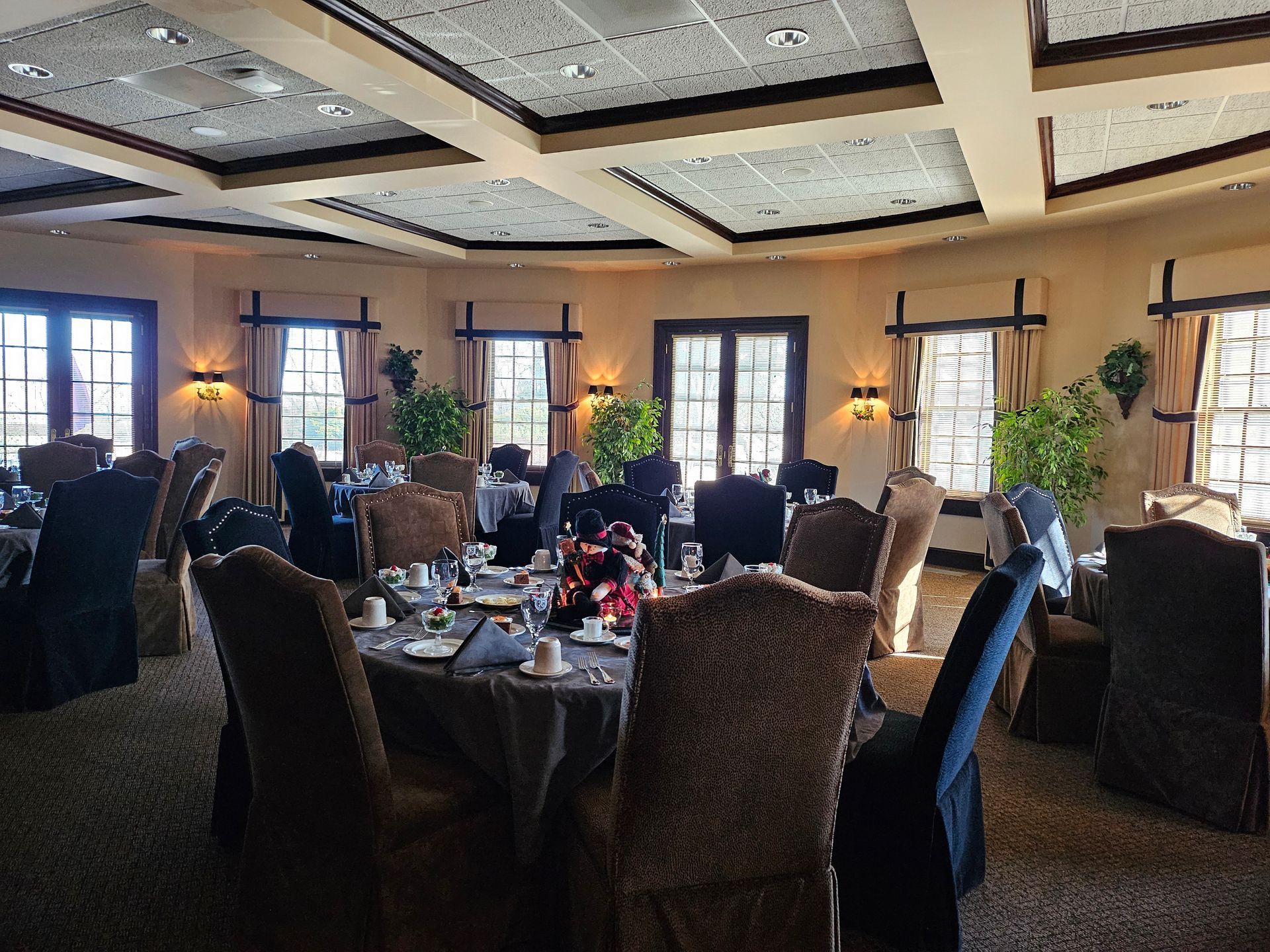 A dining room with round tables, dark upholstered chairs, large windows with brown drapes, and a decorative grid ceiling.