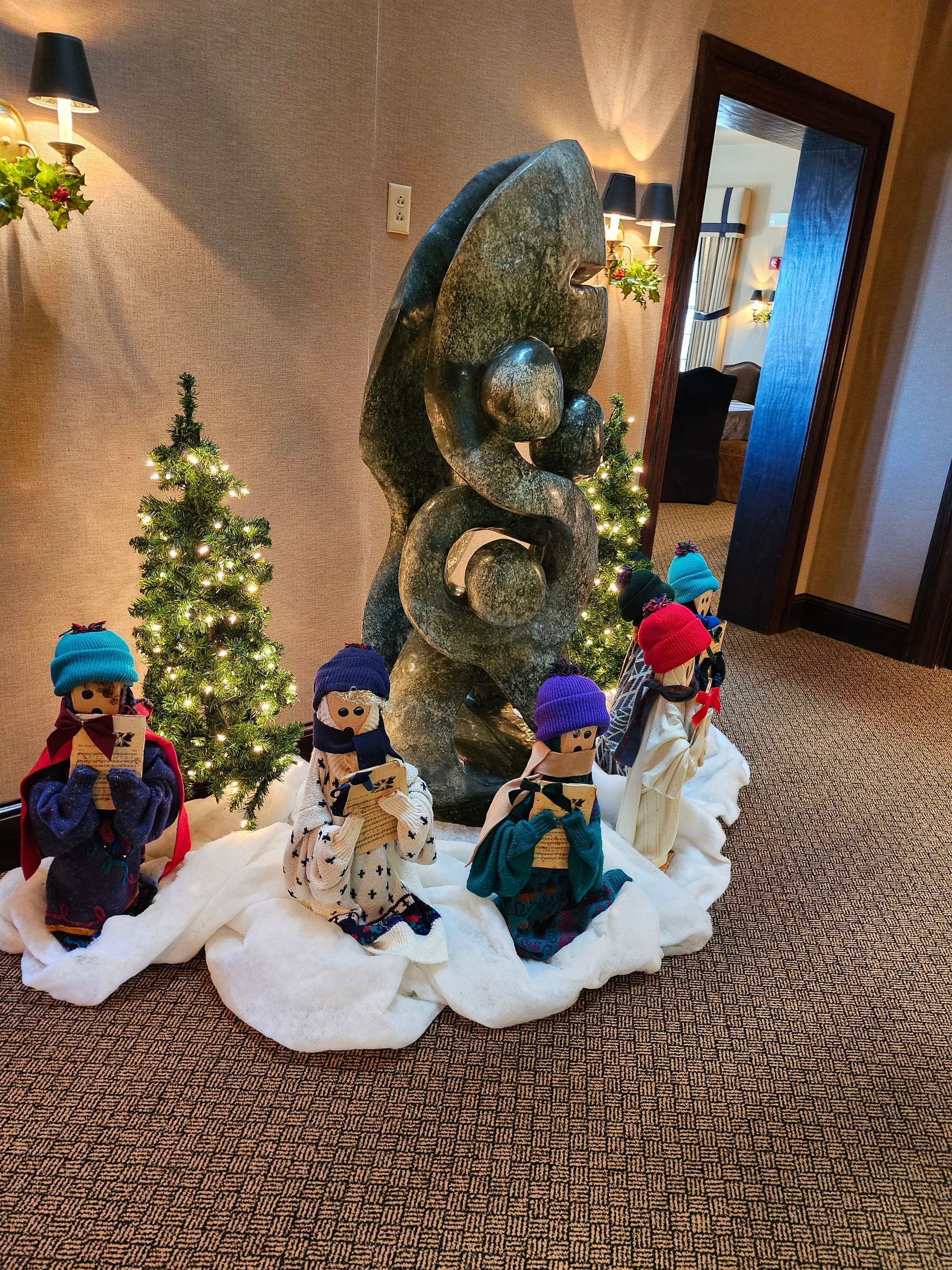 A holiday display features small figurines dressed in colorful hats and outfits arranged on artificial snow near trees.