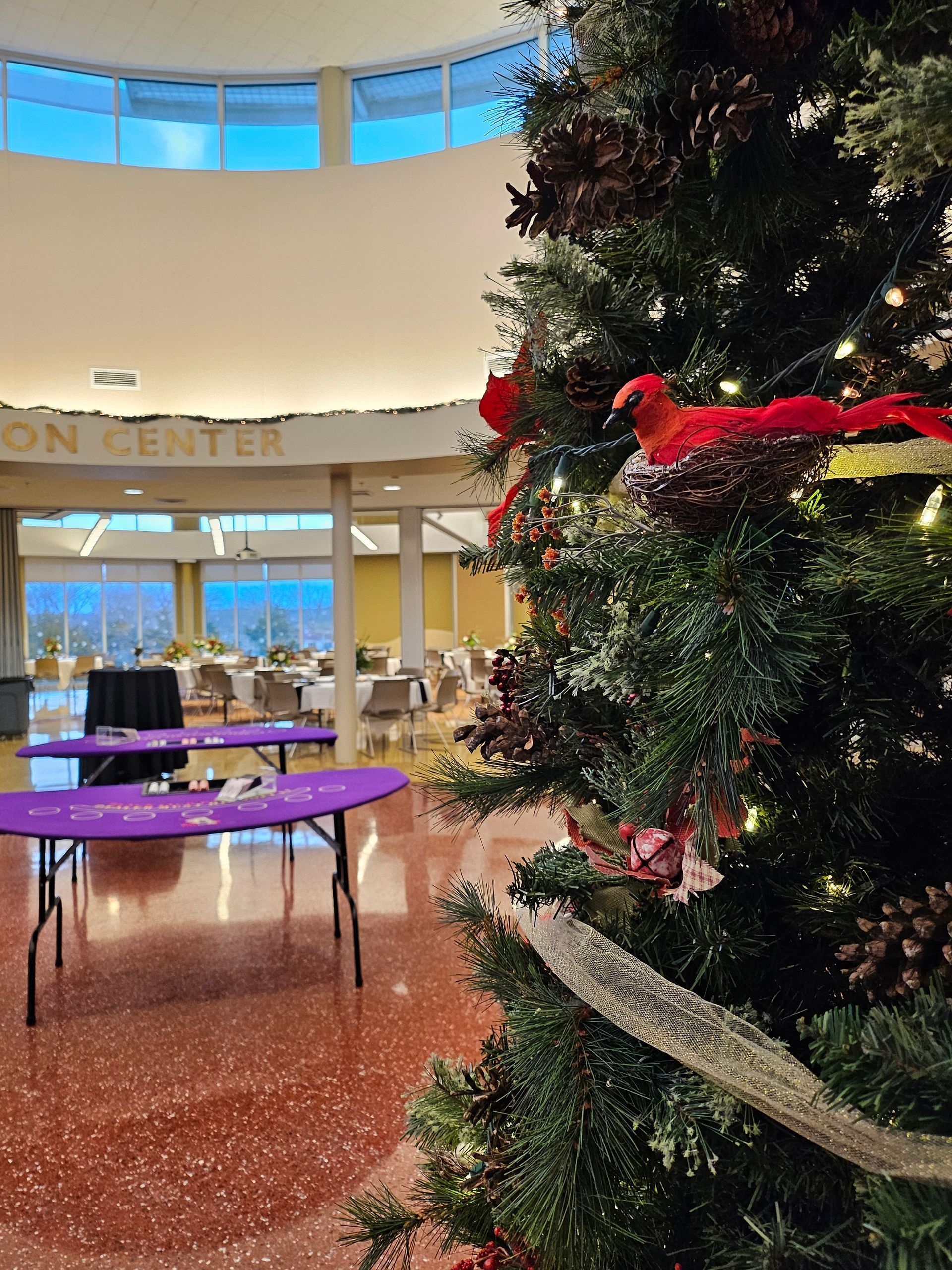 A decorated Christmas tree stands in a bright atrium, with purple-covered event tables visible in the background.