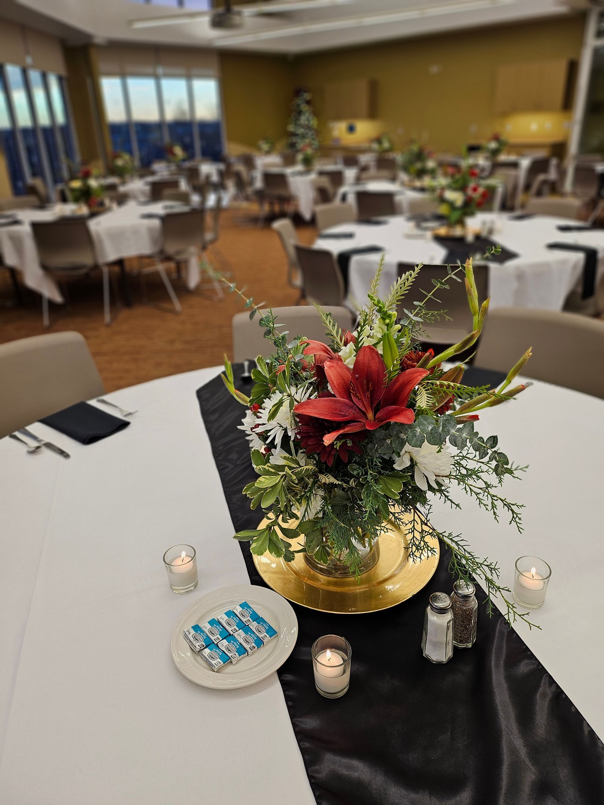 A floral centerpiece with red lilies on a table with a black runner, candles, and white tablecloths in a banquet hall.