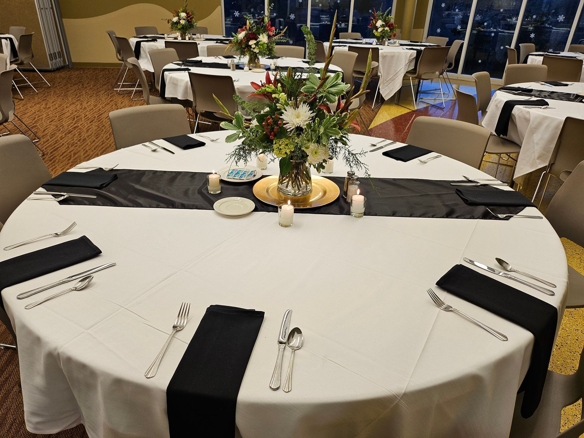 A round dining table at an event, set with a white tablecloth, black runner, floral centerpiece, and black napkins.