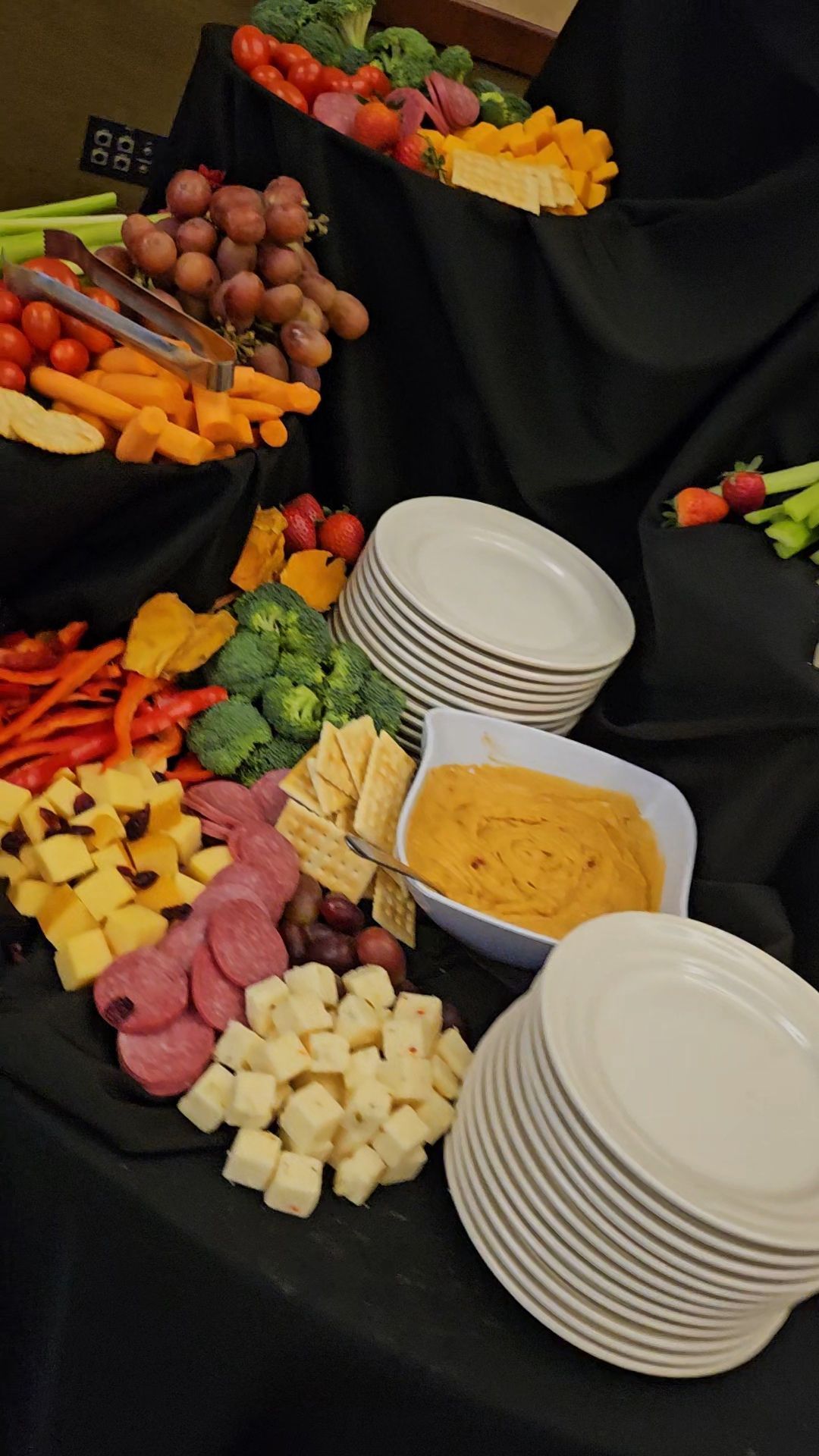 A tiered buffet table with various cheeses, meats, crackers, fresh vegetables, grapes, and a bowl of dip with stacked plates.