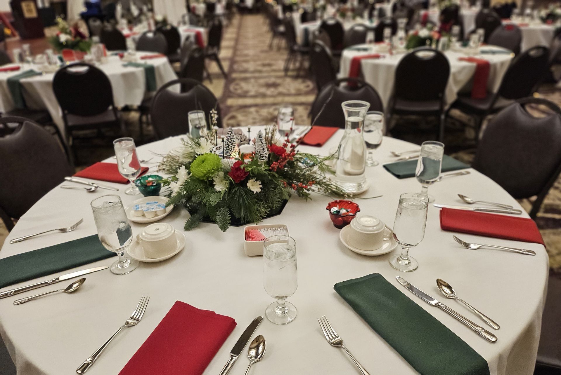 A round table at a banquet hall set for dinner with a floral centerpiece, white tablecloths, and red and green napkins.