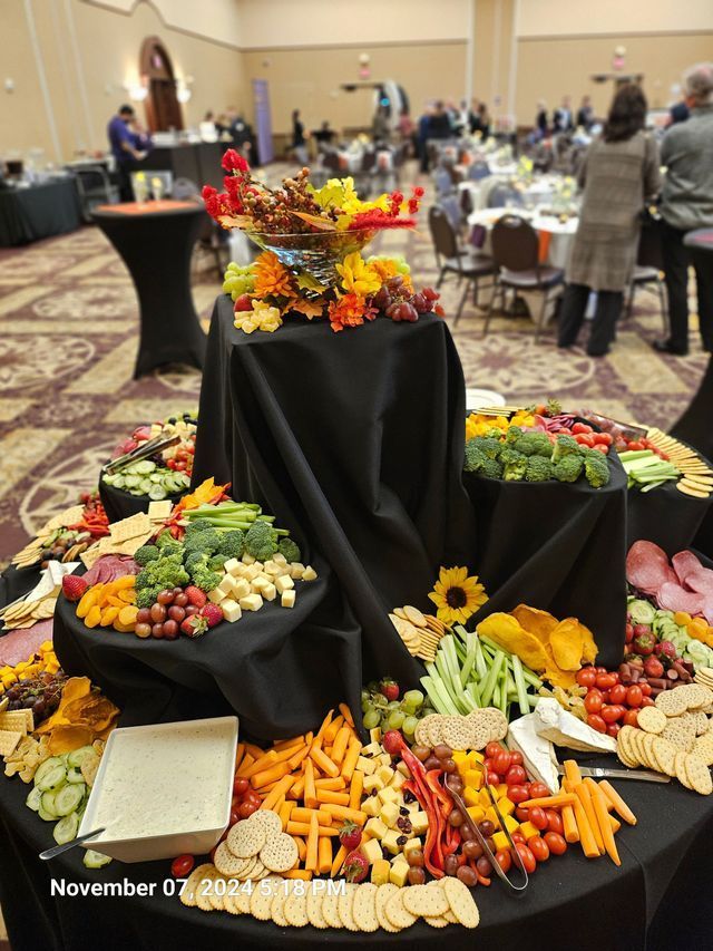 A tiered display of charcuterie, vegetables, fruit, crackers, and dip on a black-clothed table in a ballroom setting.