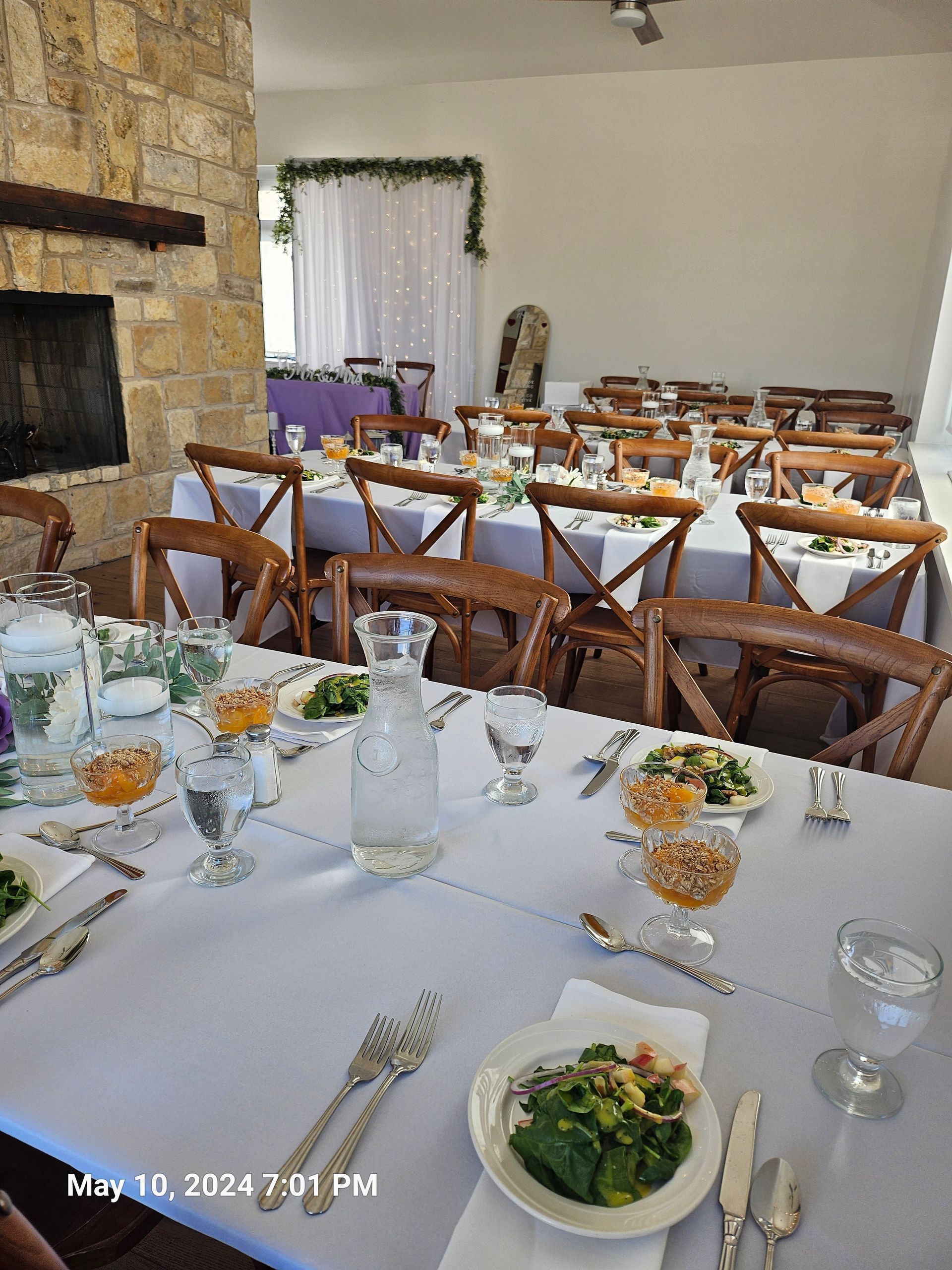 Tables set for a meal in a bright room with stone walls, wooden chairs, and a white curtain backdrop.