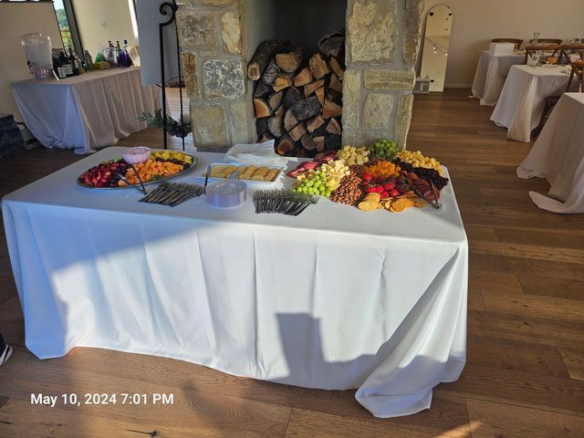A white-clothed table holding fruit, cheese, and cracker platters in a room with wood flooring and a stone fireplace.