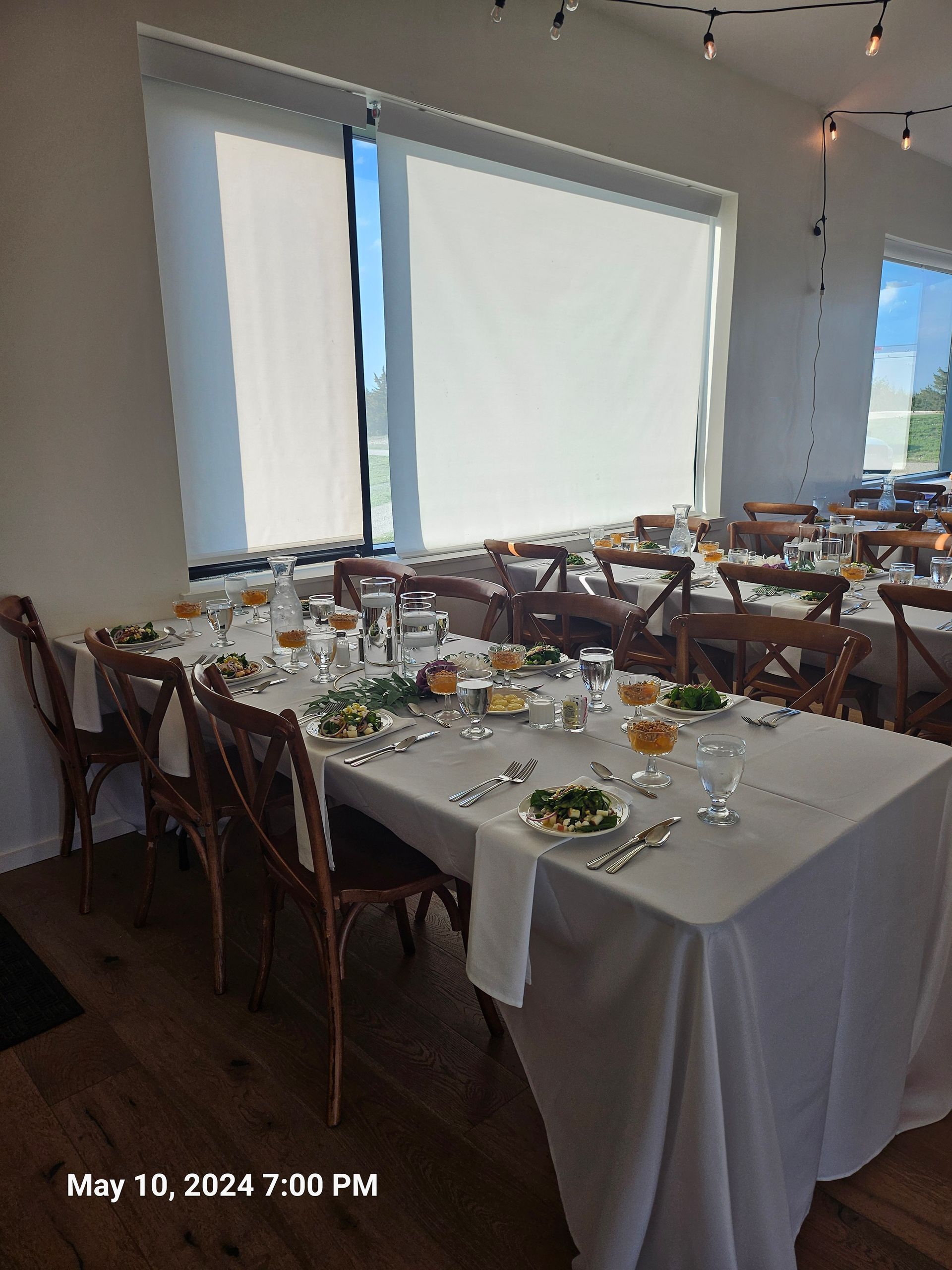 A long table set for a formal dining event with white linens, wooden cross-back chairs, and plates of salad in a hall.