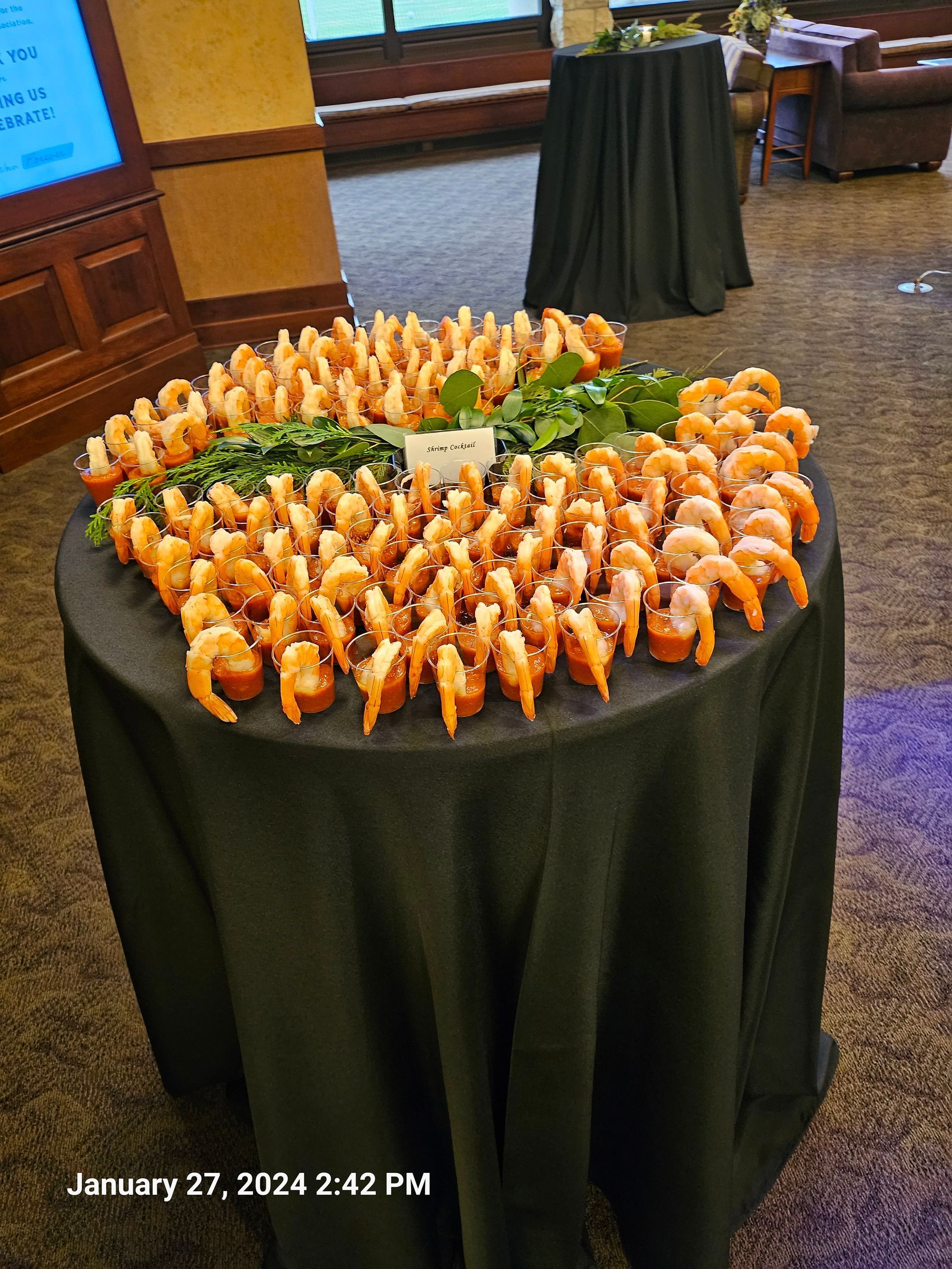 A round table with a black cloth holds a large circular display of shrimp cocktail shooters with green garnish.