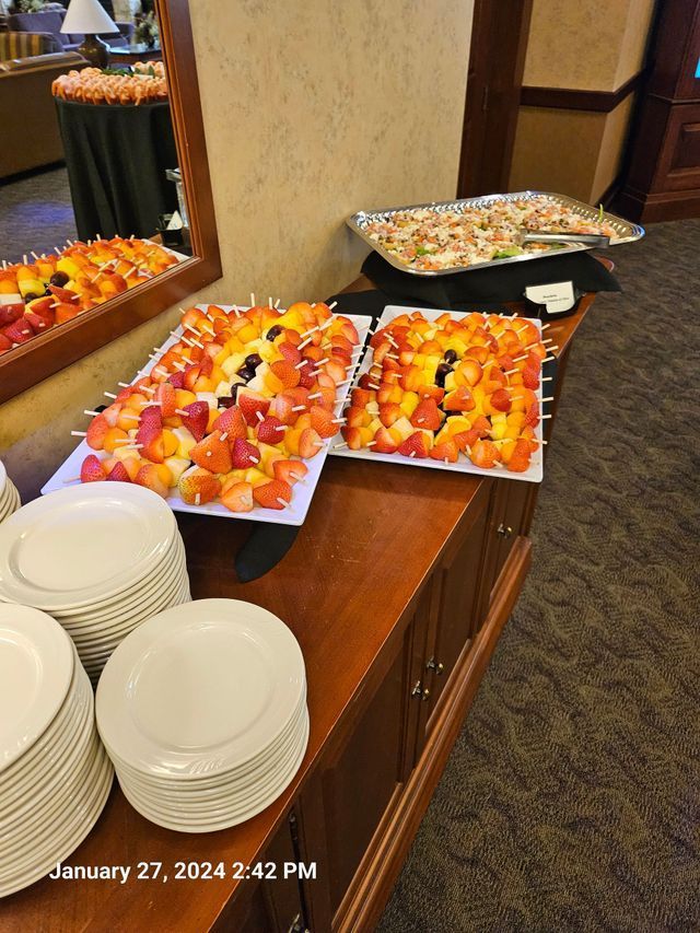 A buffet table with stacks of white plates, two platters of fruit skewers, and a tray of appetizers in a room setting.