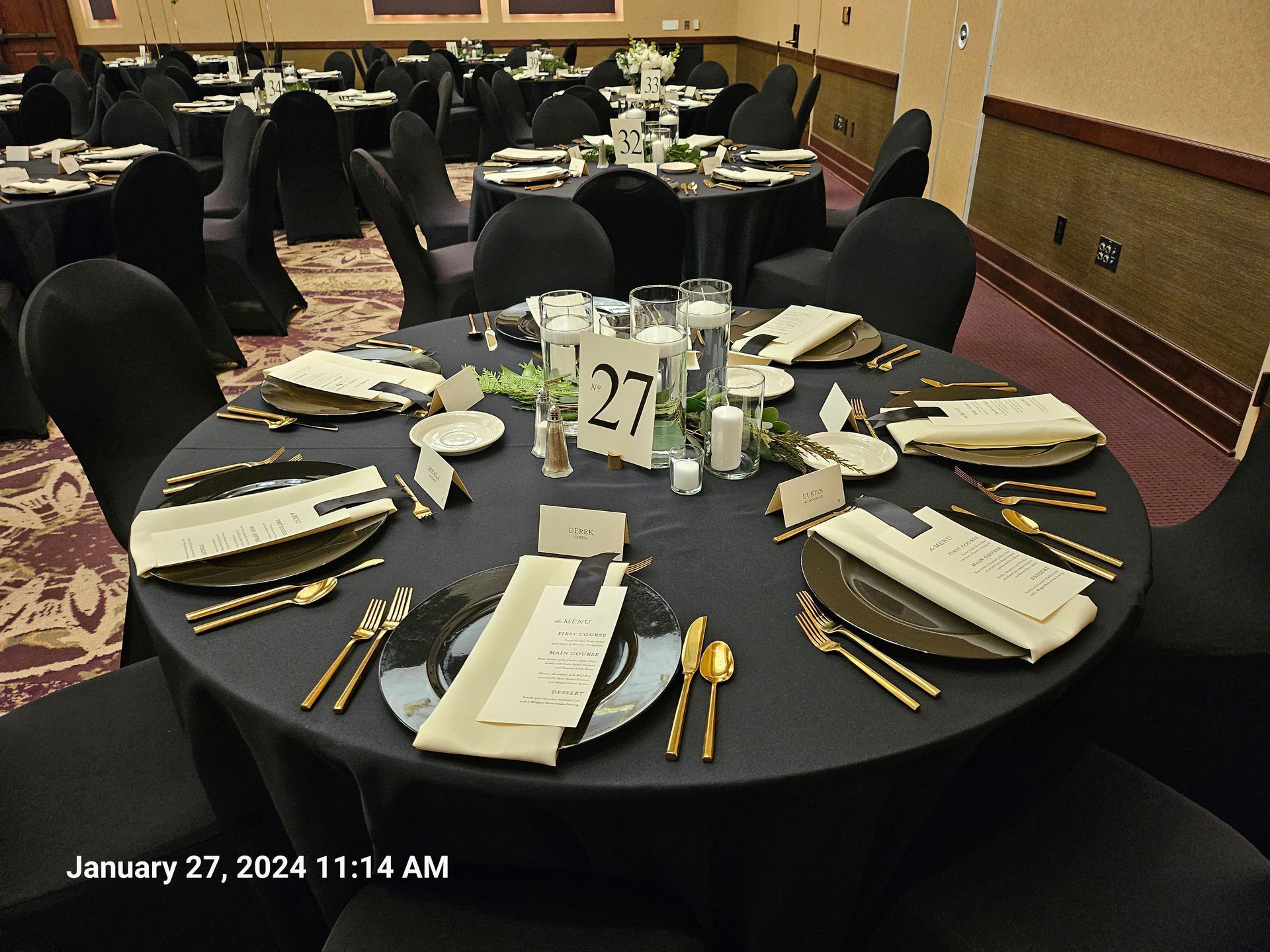 A round banquet table set for a formal dinner with black linens, gold cutlery, and a number 27 centerpiece.