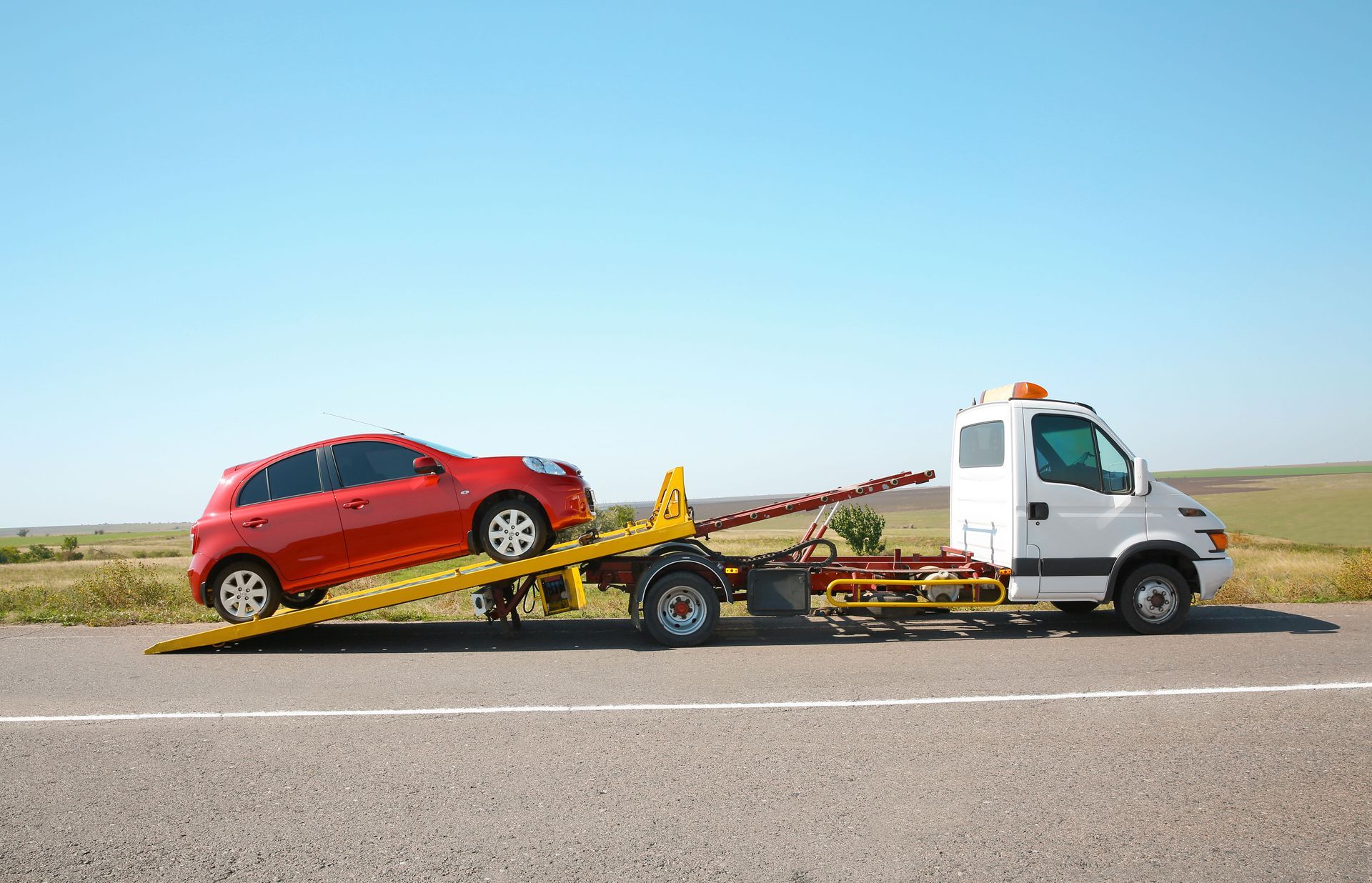 Red car being towed on a flatbed tow truck on a rural road under a clear sky.