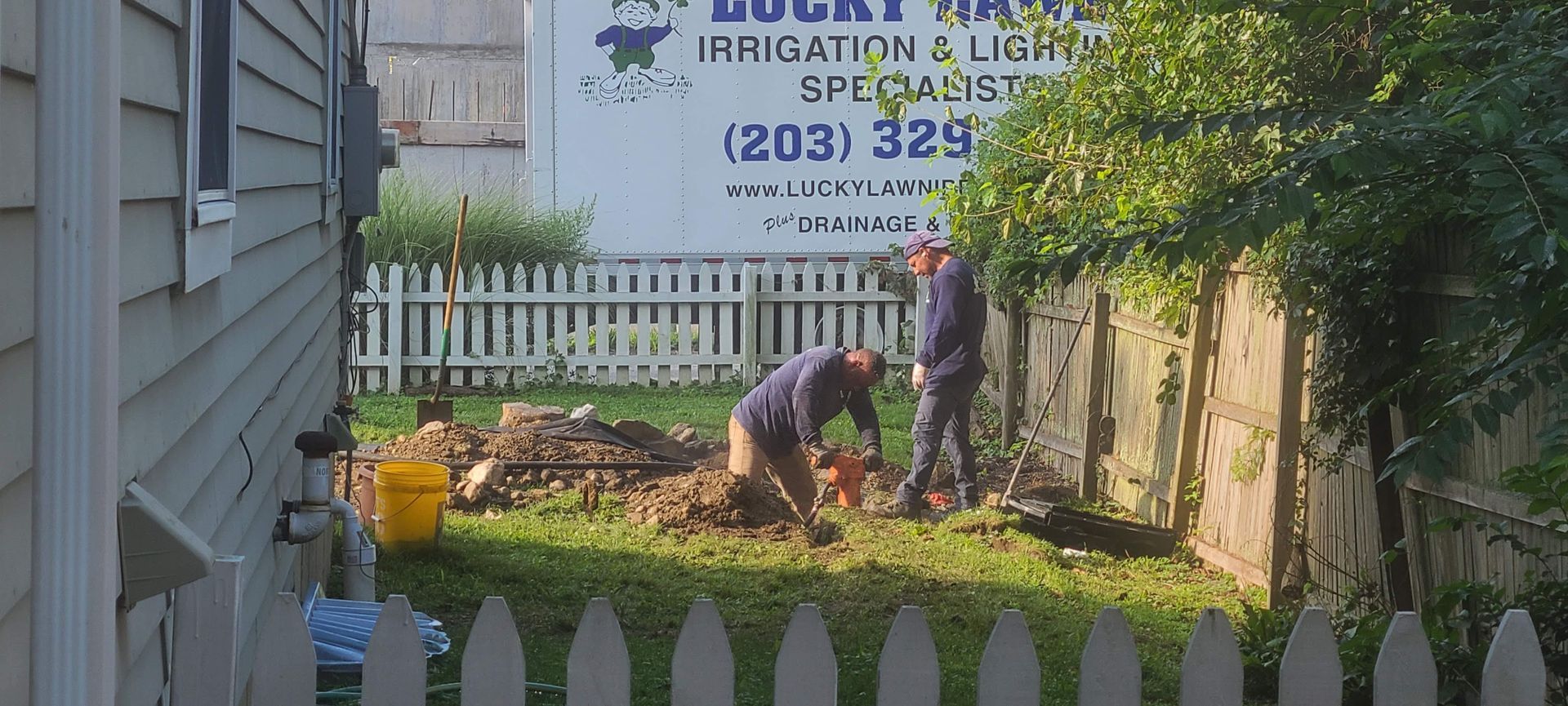 Two men are digging in a yard in front of a sign that says lucky irrigation & light