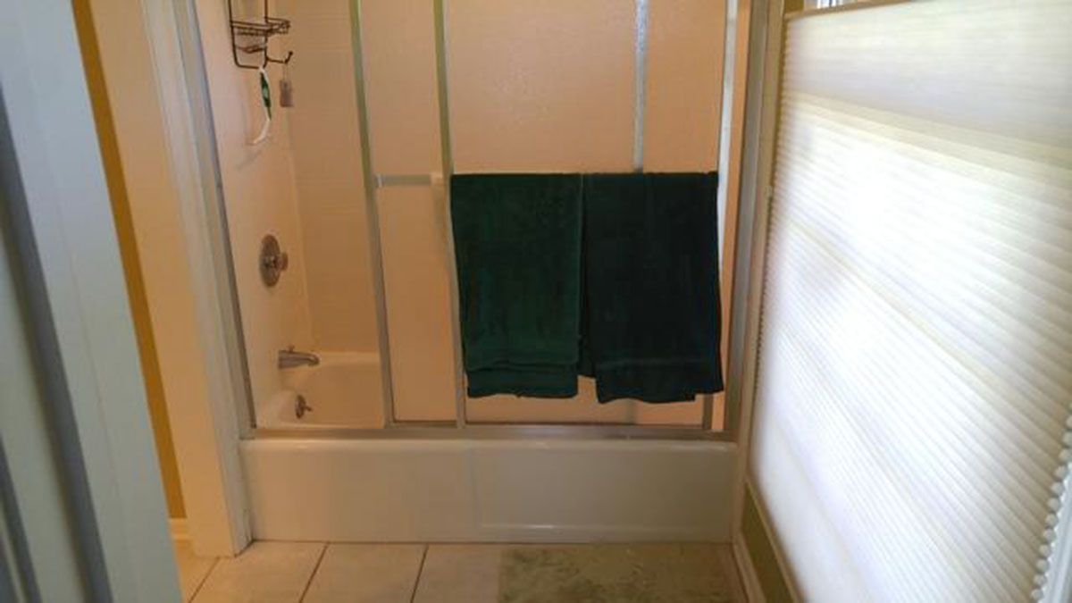 Bathroom with shower, green towels hanging inside, and a window with blinds on the right