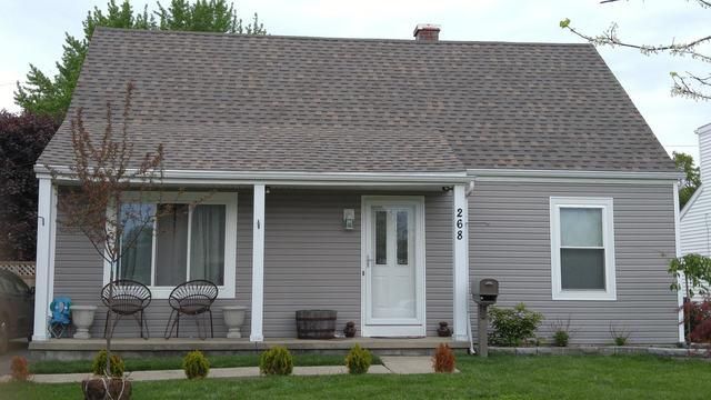 Gray house with a front porch, white door and window frames. Green lawn and a few small bushes.