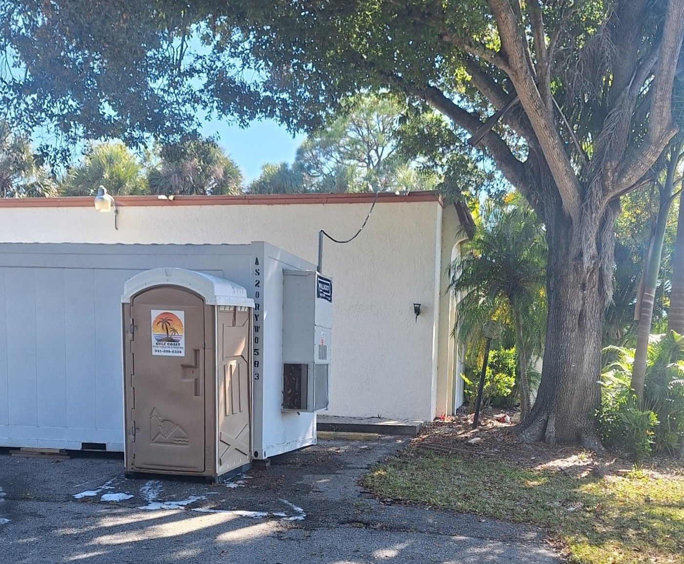 Portable toilet beside a small white building under trees on a sunny day