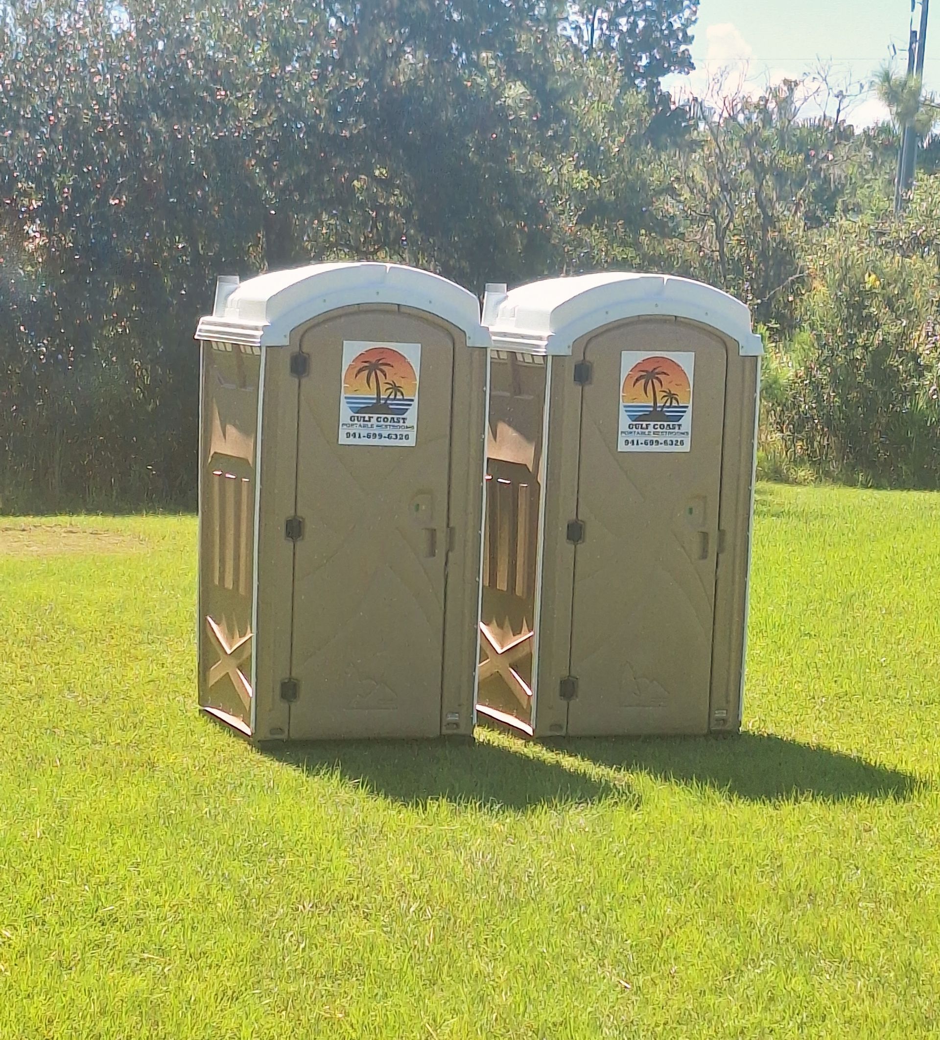 Two beige portable toilets standing side by side on a grassy field in sunlight.
