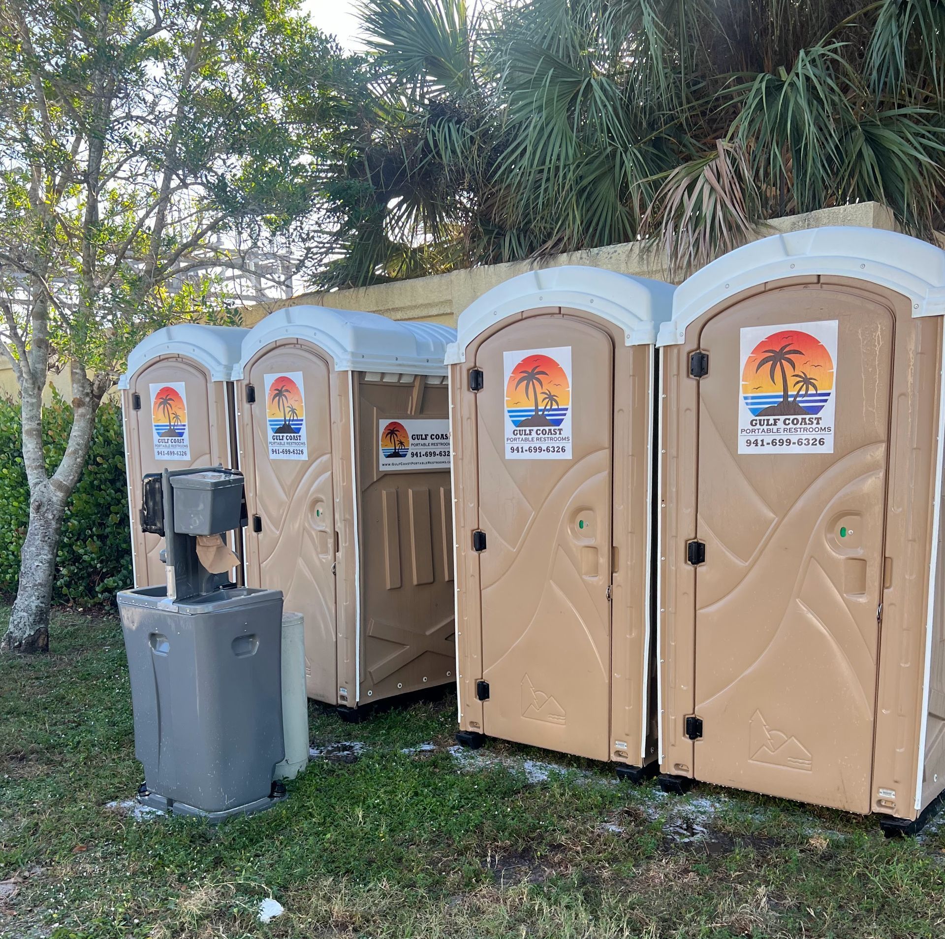 Three tan portable toilets beside a gray handwashing station in a wooded outdoor area.