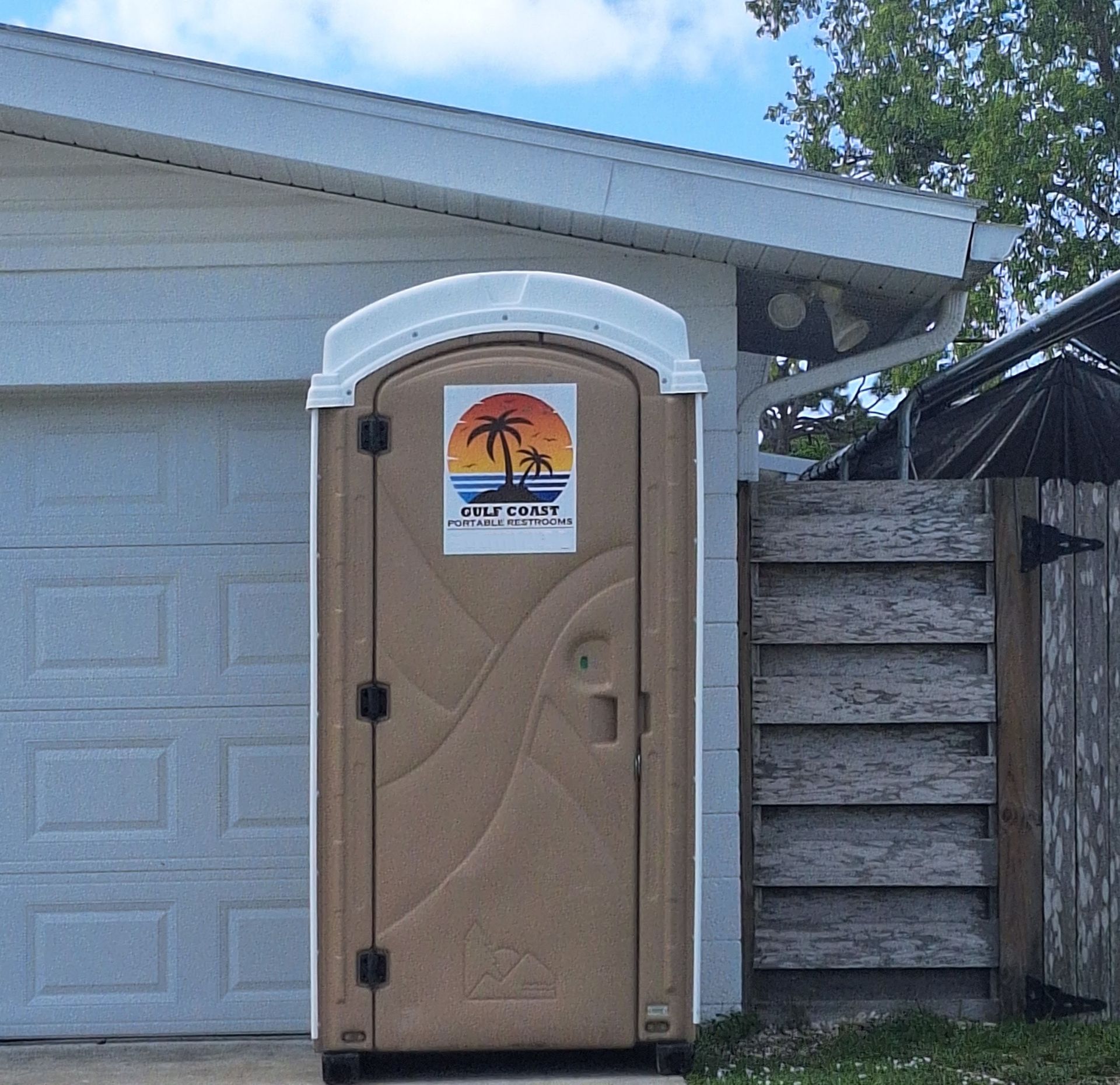 Portable toilet beside a garage, with tan door and orange sign under a white canopy