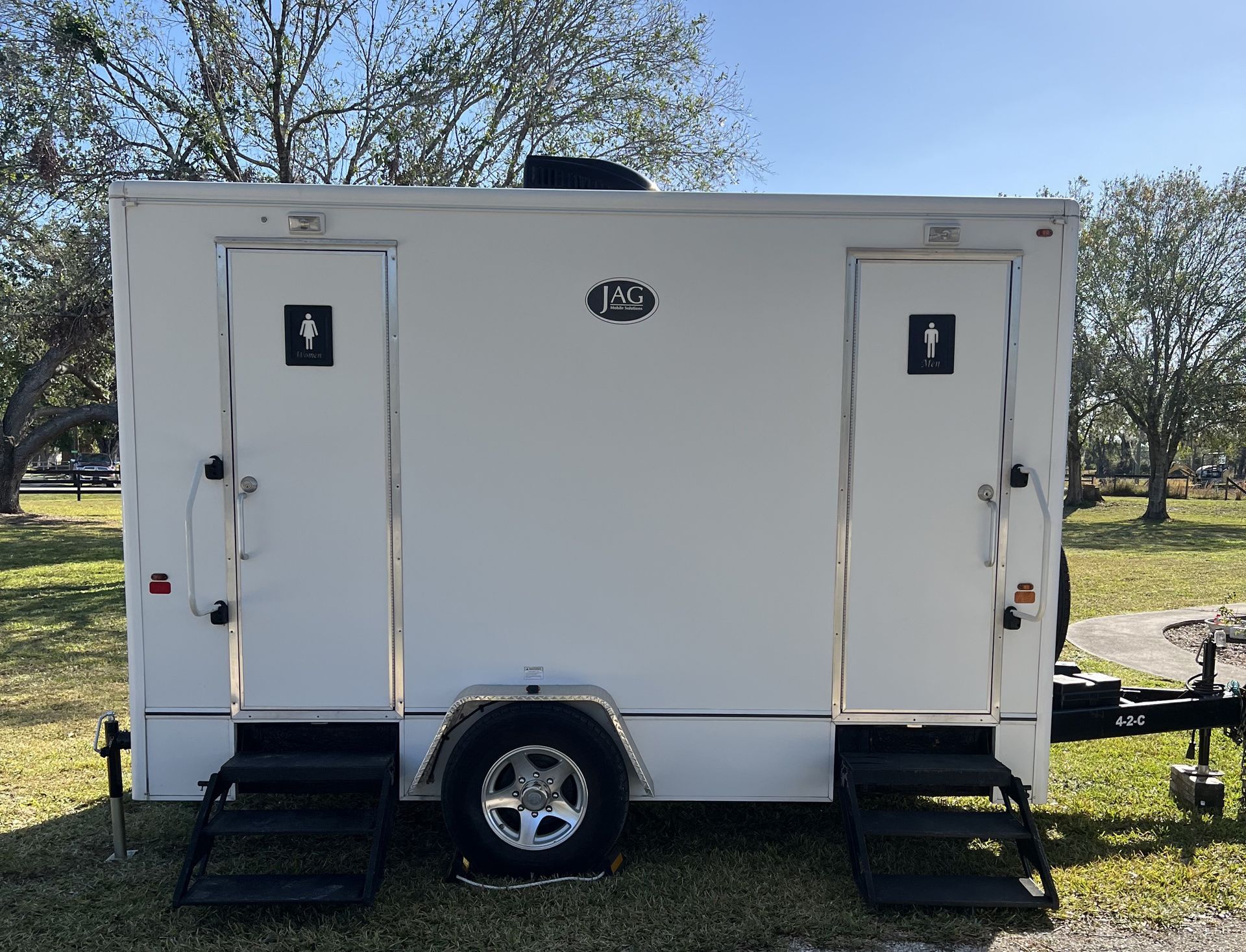 White trailer with two side doors, steps, and a logo, parked on grass with trees in the background.